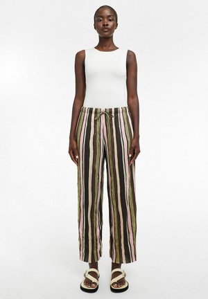 Femme debout portant un haut blanc sans manches, un pantalon ample rayé vert, rose, noir et blanc, et des sandales blanches sur fond blanc uni.