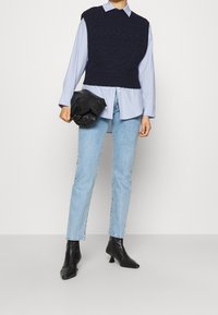 Woman wearing black knitted vest over light blue striped shirt, light blue jeans, black ankle boots, holding a black clutch bag.