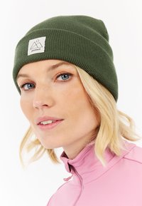 Green knitted beanie with a fold-over cuff and a square logo tag. The subject wears a pink high-collar top, with soft texture and lighter hair.