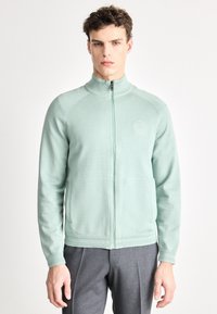 Ελαφρύ πράσινο ζακέτα με φερμουάρ σε μέντα, με ψηλό γιακά, ριμπ μανσέτες, πλαϊνές τσέπες και διακριτικό λογότυπο στο στήθος. Μαλακό υφασμάτινο υλικό.