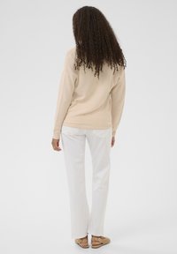 Pull en maille beige à manches longues, associé à un pantalon blanc à jambes droites et des sandales tressées couleur tan. Cheveux coiffés en boucles lâches.