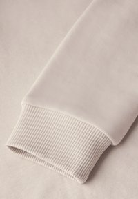 Poignet de sweat-shirt beige clair avec une texture côtelée. Tissu lisse, forme arrondie et légère brillance, mettant l'accent sur le confort et l'ajustement.