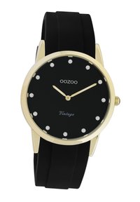 Montre noire avec un boîtier rond en or, marqueurs d'heure en cristal et aiguilles dorées. Dispose d'un bracelet en silicone noir. Le texte indique "Vintage".