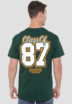 Mann trägt ein dunkelgrünes T-Shirt mit dem Aufdruck "Class of 87" und "Stranger Things" auf der Rückseite, steht mit dem Rücken zum Betrachter vor einem schlichten Hintergrund.