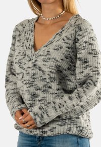 Suéter jaspeado en gris y negro con escote en V, mangas largas y puños acanalados. Textura de punto con un corte holgado. Combinado con jeans azules.