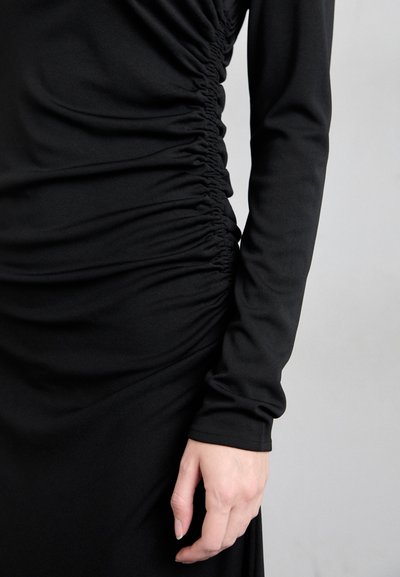Robe noire à manches longues avec fronces sur le côté, réalisée en tissu lisse. Présente une silhouette ajustée et un col rond.