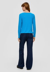 Kobieta stojąca tyłem, ubrana w jasnoniebieską bluzkę z długim rękawem, ciemnoniebieskie spodnie z szerokimi nogawkami i czarne buty na jednolitym szarym tle.