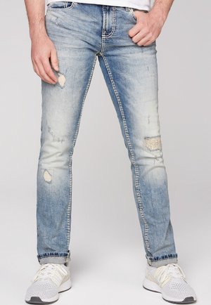 Uomo che indossa jeans azzurri consumati e sneakers bianche, in piedi con le mani vicino alle tasche su sfondo grigio.
