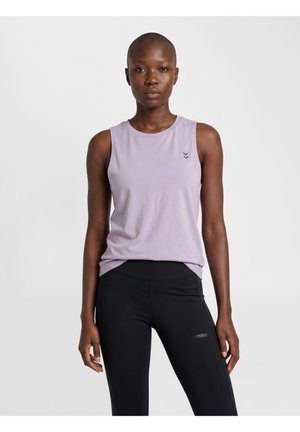 Kvinde med kort hår iført en ærmeløs, lyse lilla top og sorte tætsiddende leggings, stående mod en ensfarvet hvid baggrund.