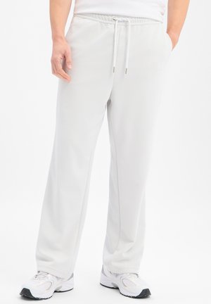 Person, die weiße, locker sitzende Jogginghosen mit Kordelzugbund und weiße Sportschuhe trägt, steht mit einer Hand in der Tasche.