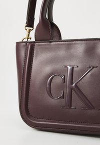 Borsa in pelle bordeaux con logo 'CK' impresso, design strutturato e una singola tracolla con accenti in metallo dorato.