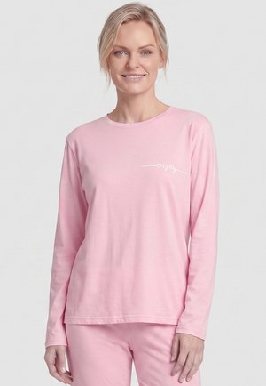 Junge Frau mit blondem Haar, die ein hellrosa langärmliges Oberteil und eine Hose mit dem Wort "enjoy" auf der Brust trägt, lächelnd und stehend.