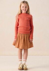 Koraal gebreide top met lange mouwen en een klein blauw embleem, gecombineerd met een bruine rok met oranje bloemenborduursels en beige sneakers.