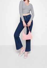 Žena v šedém zkráceném svetru a tmavě modrých džínech, která drží růžovou tkanou kabelku a má na sobě růžové tenisky, na bílém pozadí.