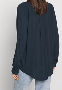 Donna che indossa una camicia a maniche lunghe blu scuro e jeans azzurri, vista da dietro su uno sfondo semplice.