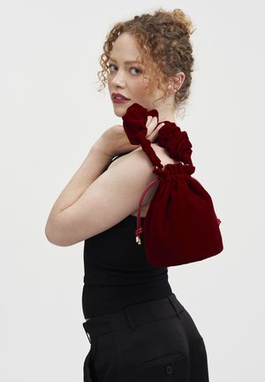 Bolsa de drawstring de terciopelo rojo con correas de tela recogidas y acentos de hardware dorado. Forma redondeada, textura suave, perfecta para un uso casual.