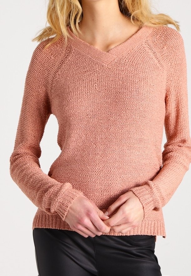 Femme portant un pull en maille rose pâle à col en V avec manches longues, assorti à un pantalon noir, les mains jointes à la taille.