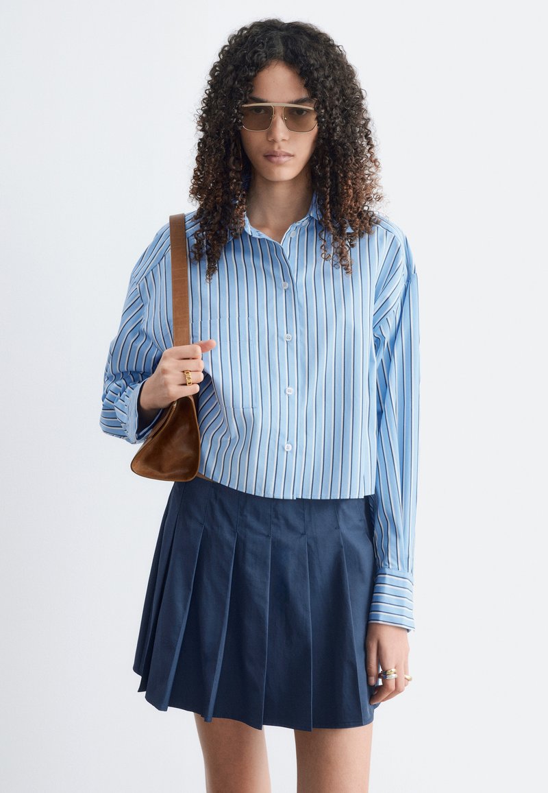 Femme aux cheveux bouclés portant des lunettes de soleil, une chemise à rayures bleues boutonnée, une jupe plissée bleu marine, et tenant un sac à bandoulière marron.
