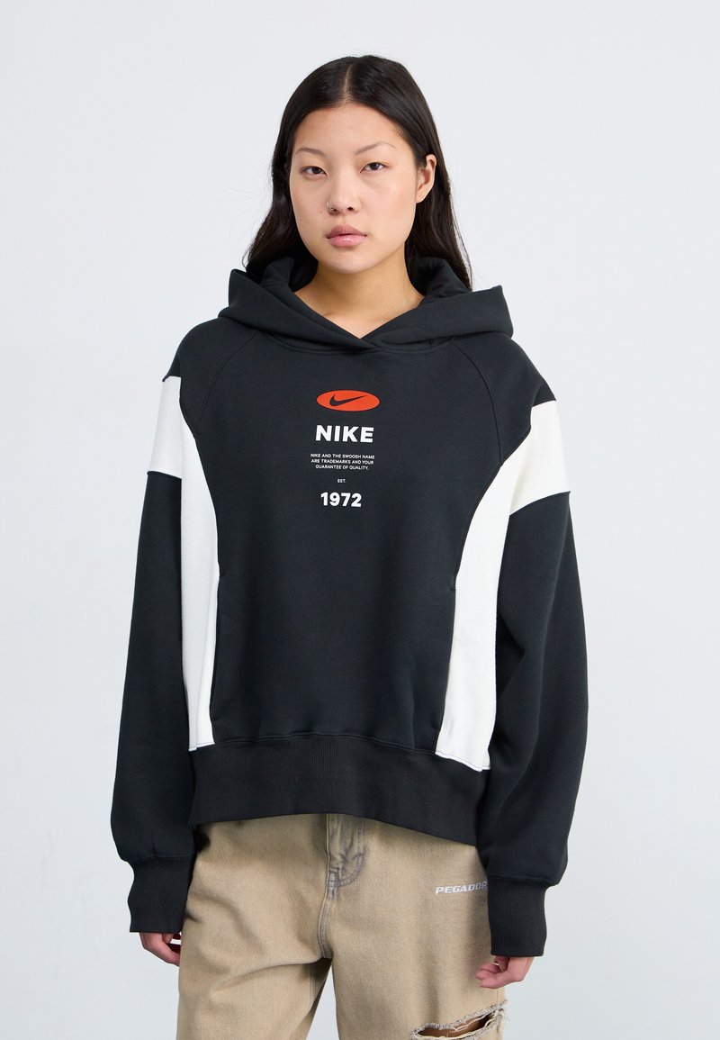 Hoodie noir avec des accents latéraux blancs, arborant un logo Nike orange en évidence et du texte. Fabriqué en tissu doux avec une coupe décontractée.
