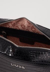 Černá kožená taška s krokodýlím vzorem a stříbrným zipem, otevřená a odhalující světle růžovou podšívku s vzorem a vnitřní kapsou na zip.