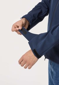 Main ajustant la manche d'une veste bleu marine avec sangle en velcro, fermeture éclair visible, personne portant un jean bleu, isolée sur un fond clair.