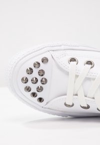 White sneaker with flat laces, rubber sole, and metal studs arranged on the side. Smooth texture, rounded toe design, and subtle stitching details.