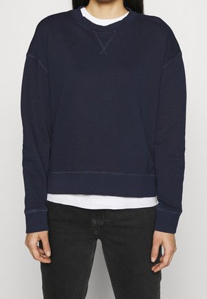 Person trägt einen marineblauen Pullover mit langen Ärmeln über einem weißen Hemd, kombiniert mit schwarzen Jeans, steht vor einem einfarbigen Hintergrund.