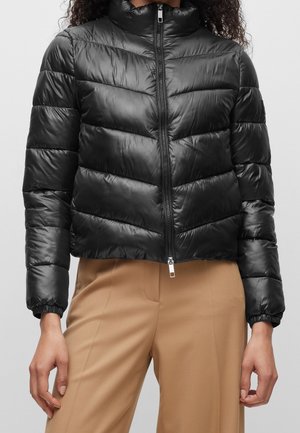 Zwarte doorgestikte pufferjas met rits aan de voorkant, gedragen met beige broek met hoge taille, handen ontspannen langs het lichaam.