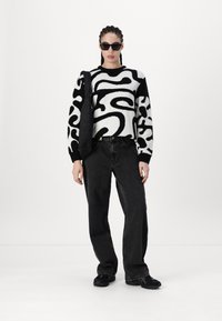 Gebreid zwart-wit trui met abstract patroon, gecombineerd met zwarte wijde jeans en zwarte sneakers. Model draagt kattenoogzonnebril.