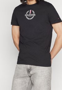 Musta puuvillainen T-paita, jossa on pyöreä kaula-aukko, lyhyet hihat ja valkoinen logo, jossa on laakerinlehdet ja punainen korostusviiva.