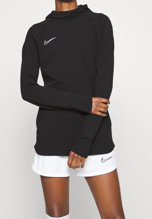 Osoba ubrana w czarną bluzę z kapturem Nike z długim rękawem oraz białe sportowe spodenki Nike, stojąca na jednolitym jasnym tle.