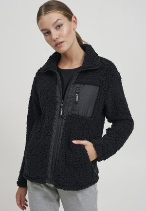 Black zip-up jacket with a fuzzy texture, featuring a smooth nylon front pocket and side pockets. High collar and elastic cuffs.