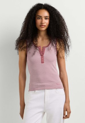 Jonge vrouw met lang krullend haar, die een mouwloos roze gestreept topje en een witte broek draagt, staand tegen een effen lichte achtergrond.