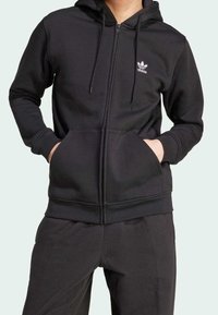 Černá zipová mikina vyrobená z jemné tkaniny. Obsahuje přední kapsu, kapuci na šňůrku a malé bílé logo Adidas na levé hrudi.