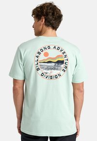 Mėtų spalvos medvilninis marškinėliai su apvaliu piešiniu, kuriame yra saulė ir kraštovaizdis, ir tekstas "Billabong Adventure Division". Trumpomis rankovėmis, laisvas kirpimas.