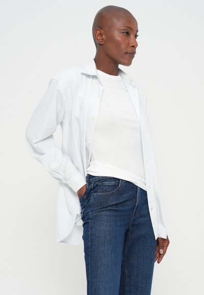 Persona con cabeza rapada, usando una camiseta blanca, camisa abierta de rayas claras y jeans azules, de pie con una mano en el bolsillo contra un fondo blanco.