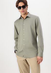 Chemise à manches longues, boutonnée, de couleur vert olive, en tissu lisse, avec un col classique et des boutons métalliques, associée à un pantalon beige clair.