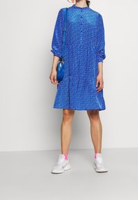 Blå blommig klänning med knäppning fram, långa ärmar och en volangkjol. Till det bärs vita sneakers och en liten blå handväska.