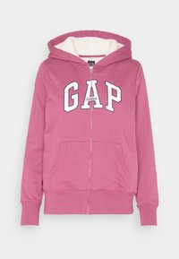Růžová zipová mikina z měkké tkaniny, s bílým nápisem v bloku "GAP" a klokankovými kapsami. Uvnitř je podšitá plyšovým materiálem.
