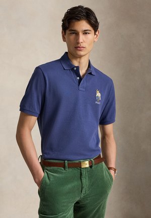 Młody mężczyzna w niebieskiej koszulce polo z logo niedźwiedzia i zielonych spodniach sztruksowych z brązowym paskiem, stojący z rękami w kieszeniach.