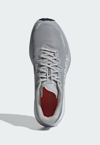 Zapatilla deportiva gris con parte superior de malla, acabado texturizado y interior rojo contrastante. Presenta cordones blancos y una marca en el costado.