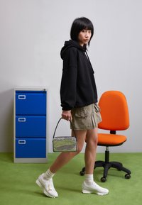 Schwarzer übergroßer Kapuzenpullover, khakifarbene Cargo-Shorts mit Taschen, weiße Sneaker und eine kleine silberne Tasche in rechteckiger Form. Grüner Teppichboden.