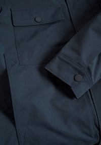 Chaqueta azul marino, hecha de una tela suave, con un bolsillo en el pecho que cuenta con un botón a presión y una manga estructurada con sutiles detalles de costura.