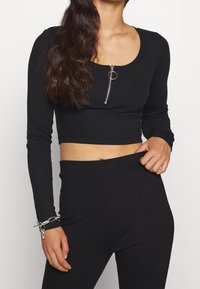 Schwarzes, geripptes Langarm-Crop-Top mit rundem Ausschnitt und einem silbernen Reißverschluss mit einem runden Zipper. Weiche Textur mit tailliertem Design.