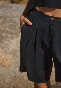 Pantalones cortos de lino negro con pliegues y cierre con un solo botón. Presentan un bolsillo lateral y una textura ligera, adecuados para clima cálido.