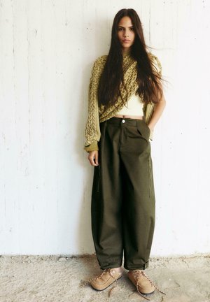 Donna con capelli lunghi e scuri che indossa un maglione lavorato a maglia grossa verde oliva, pantaloni larghi scuri e scarpe stringate color cammello, in piedi contro un muro bianco.