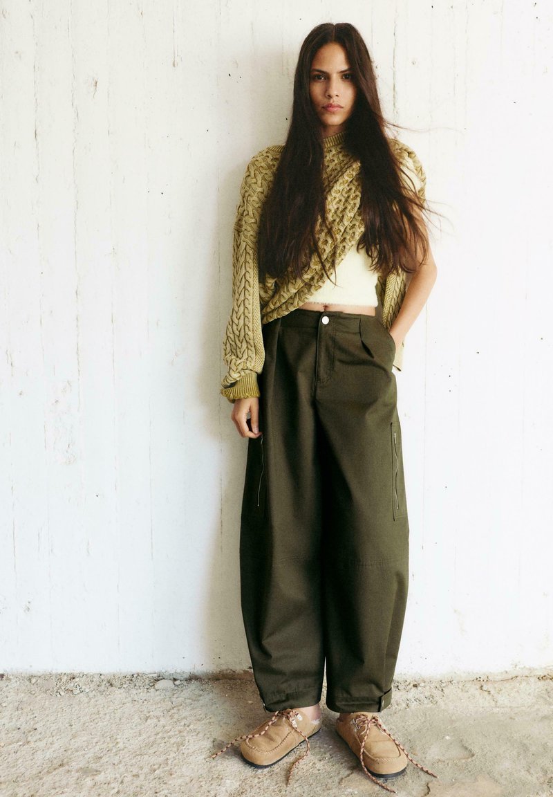 Ženska z dolgimi temnimi lasmi, oblečena v grobo pleten olivno zelen pulover, široke temne hlače in rjave čevlje z vezalkami, stoji ob belem zidu.