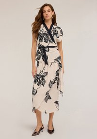 Donna che indossa un vestito avvolgente beige con grandi stampe floreali nere e una cintura nera, abbinato a tacchi metallici a punta, in piedi su uno sfondo semplice.