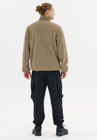 Man with hair tied in a bun wearing a brown fleece jacket and black cargo pants standing facing away on a white background.