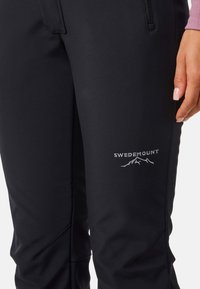 Zwarte softshell broek met een gladde textuur. Voorzien van een ritszak en een zilveren "SWEDEMOUNT" logo met een accent van een bergontwerp.